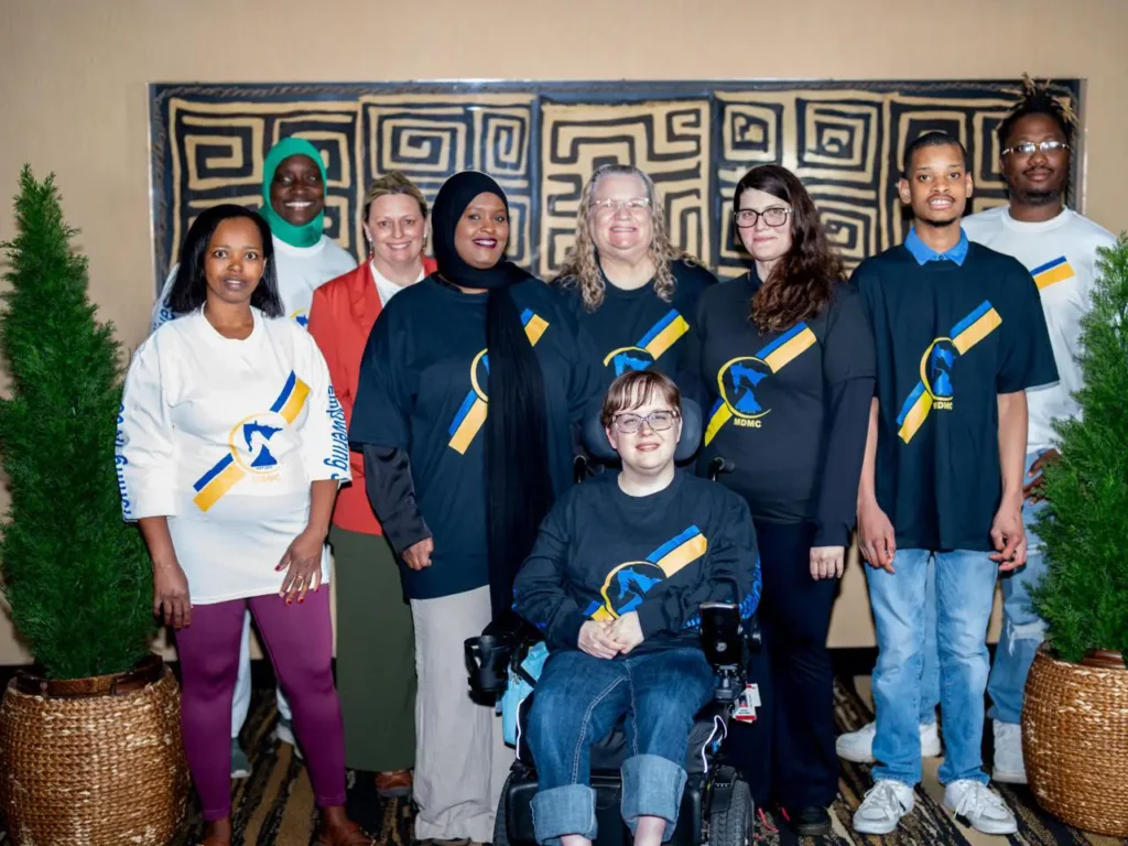 A diverse group of nine people, including one in a wheelchair, pose together indoors in front of a decorative wall, some wearing matching shirts.